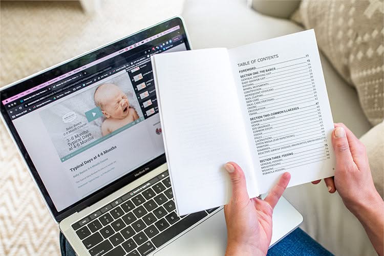 A person holds a book open to the table of contents next to a laptop displaying baby course