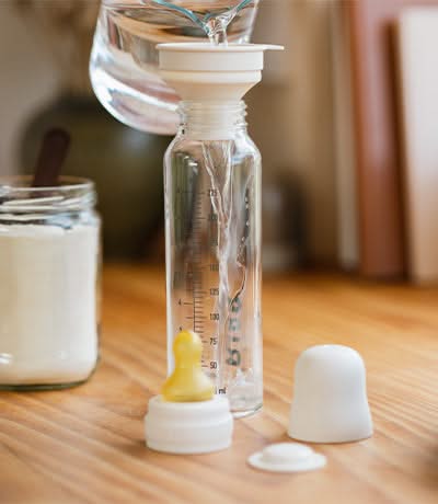 A baby bottle with a funnel on top being filled with powdered formula from a clear glass jar