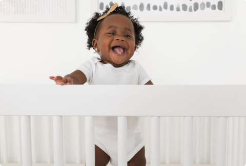 A black toddler girl is holding on to her crib and smiling
