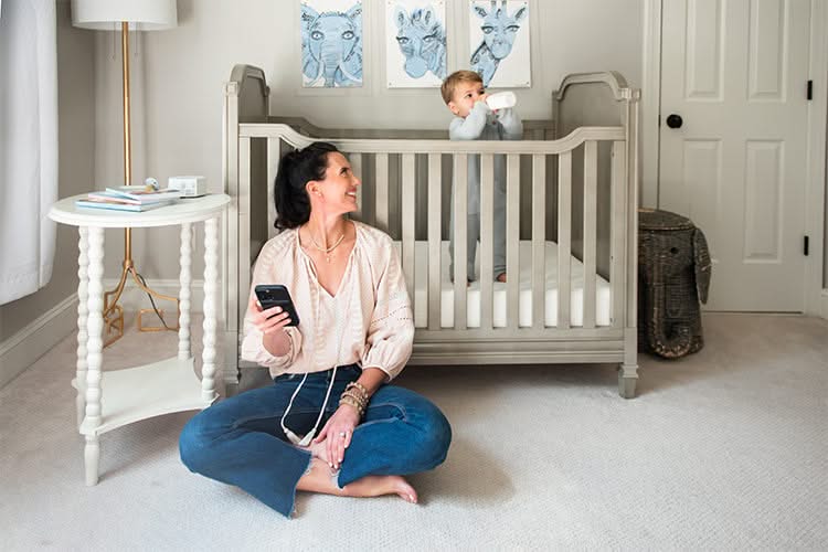 Woman sitting on the floor, smiling at her baby who stands in a crib in a well-decorated nursery
