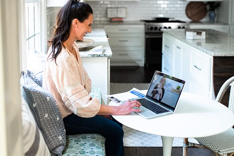 A woman smiles while using a laptop at a kitchen table, engaged in a video call displayed on her screen