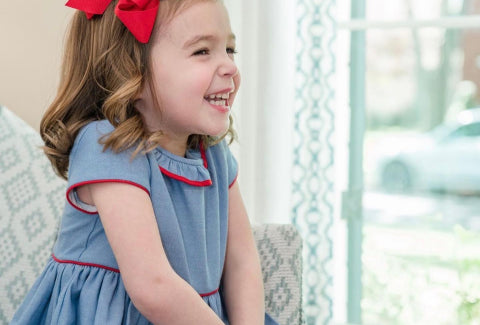 A joyful young girl with curly hair, wearing a blue dress and a red bow, laughing while sitting near a window