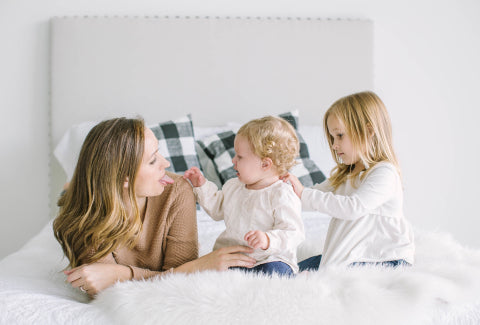 A mother lies on a white bed with her young children, a toddler and a school-aged girl