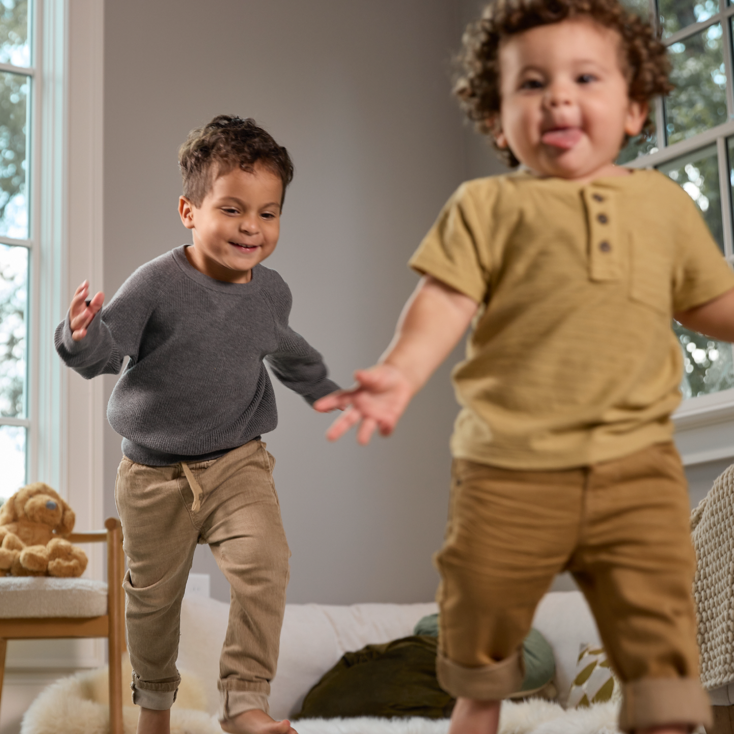 Two young boys playing together in a room with a teddy bear and furniture.
