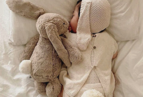 A baby in a white knitted outfit and hat sleeps on a bed, clutching a bunny plush toy