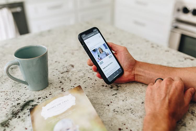 A person's hand holding a smartphone displaying a social media profile, with a coffee mug and an open magazine on a kitchen counter