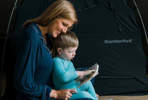 Mother holding a toddler while he reads a book, the SlumberPod logo in the background